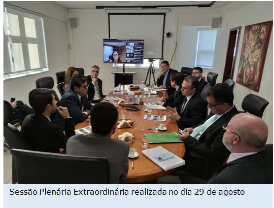 Foto horizontal de uma mesa redonda com 11 homens vestindo terno e duas mulheres uma presencial e outra online.