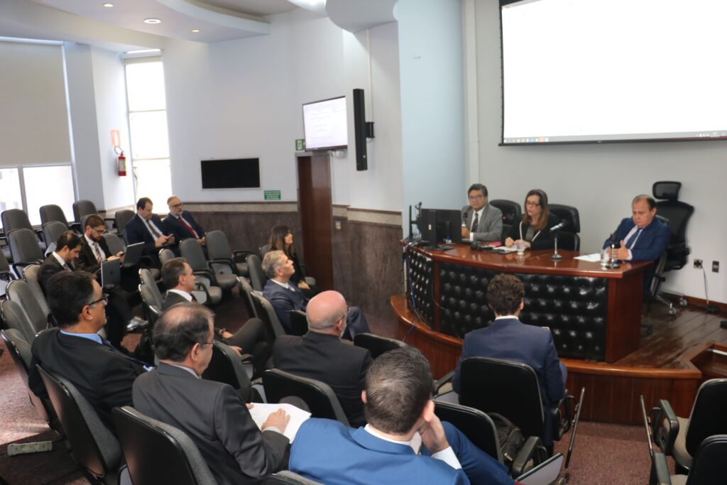 foto vertical colorida em um auditório com varias pessoas sentadas na plateia. enquanto três pessoas estão em um palco dois homens e uma mulher, sentados em cadeiras com uma mesa a frente. a mulher ao centro  esta falando ao microfone para a plateia.