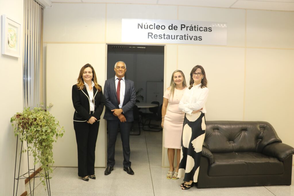 foto horizontal quatro pessoas mulher branca vestido roupas pretas e brancas, homem negro de terno e gravata, mulher branca de vestido rosa claro, mulher branca de oculos com roupas branca e preta. em frente uma sala com uma placa na porta escrito Núcleo de praticas restaurativas