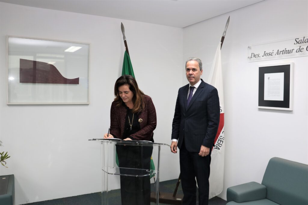 foto horizontal uma mulher e um homem em pé em uma sala. mulher assinando documento enquanto o homem observa. presidente do trf6 assina o acordo. ao lado, o presidente do TRE-MG