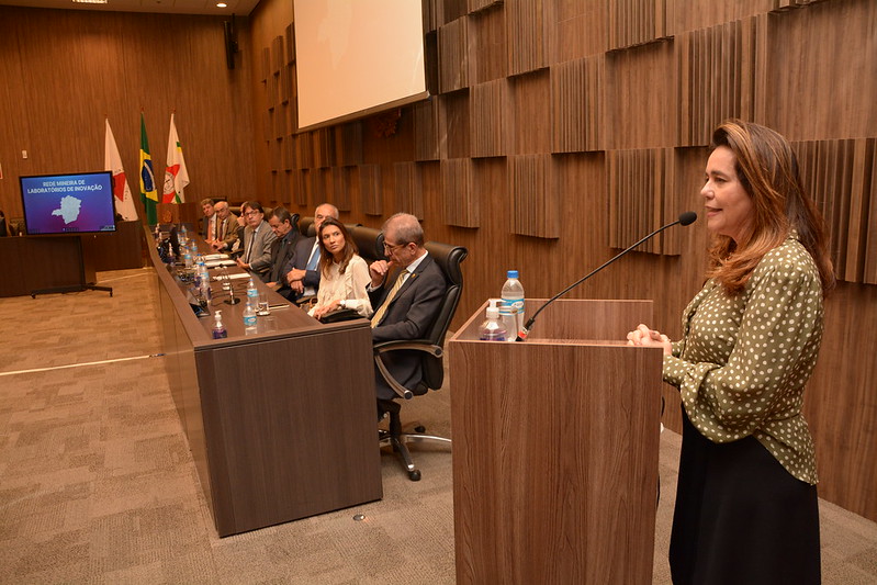 Fotografia horizontal colorida várias pessoas sentadas mesa de honra de evento uma mulher em pé falando ao microfone. Legenda: A presidente do TRF6 compôs a mesa de honra da cerimônia