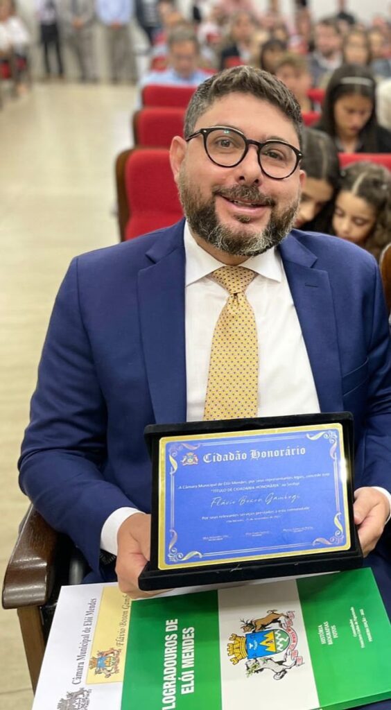 Fotografia vertical colorida homem de óculos sentado segurando placa de homenagem. Legenda: desembargador federal Flávio Boson Gambogi