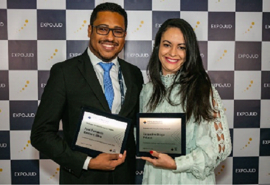 Foto colorida horizontal duas pessoas (um homem e uma mulher) em pé lado a lado, cada um segurando uma placa de homenagem.