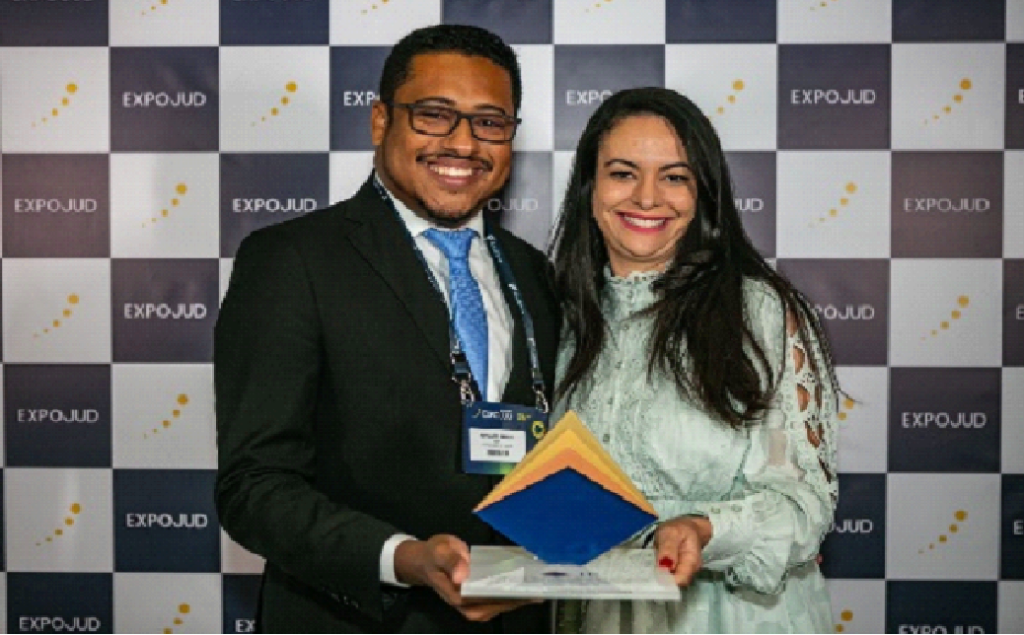 Foto colorida horizontal duas pessoas (um homem e uma mulher) em pé lado a lado segurando um troféu.