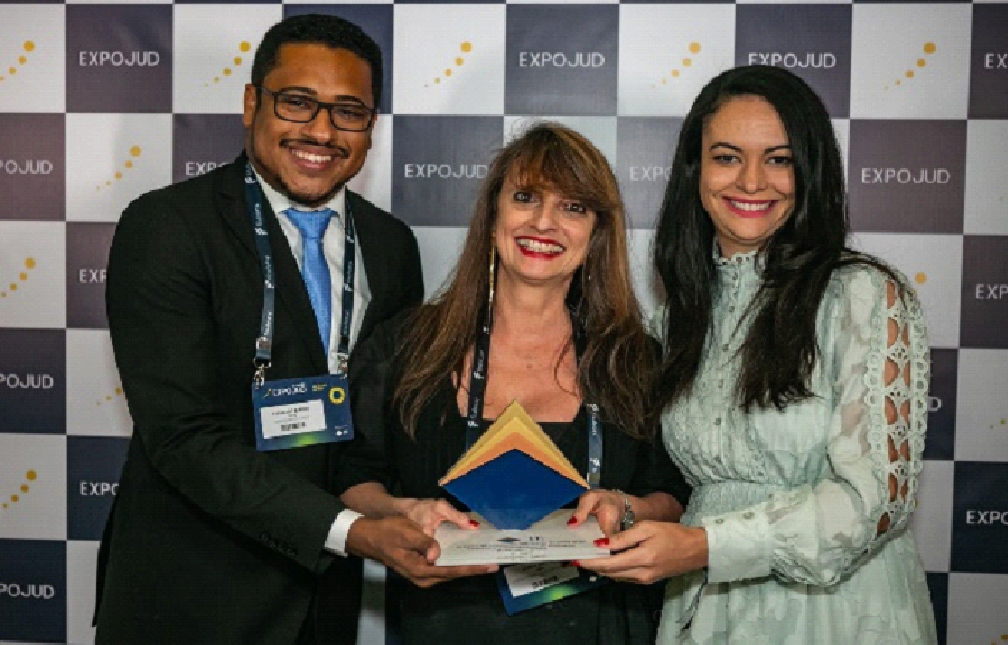 Foto colorida horizontal três pessoas (um homem e duas mulheres) em pé lado a lado segurando um troféu.