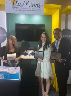 Fotografia vertical colorida duas mulheres e um homem em pé lado a lado cada um segurando uma placa de homenagem, em frente a um stand de exposição. 