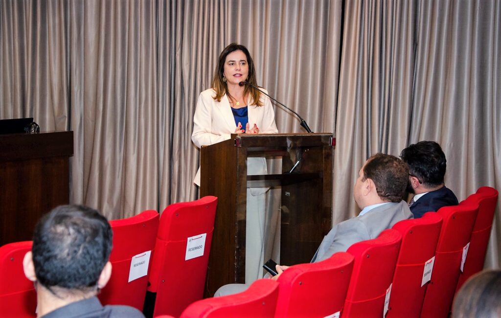 Foto horizontal colorida. Presidente do TRF6 ministra palestra para uma plateia. Ela está num púlpito de madeira escura. 