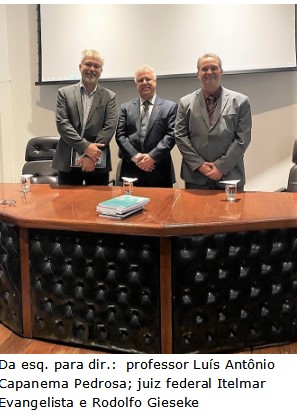 Imagem colorida vertical, com três homens em pé, atrás de uma mesa num auditório. Os três estão posando para a foto e usam roupas formais na cor cinza. 