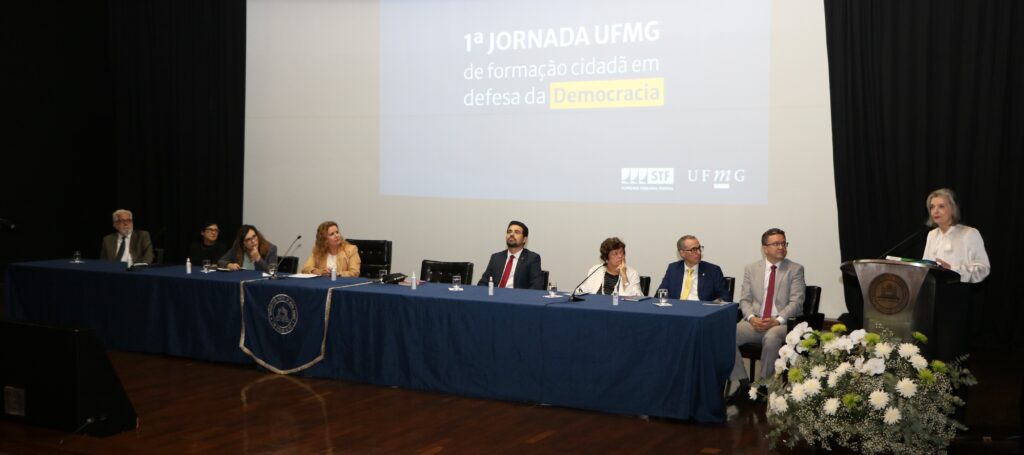 Fotografia retangular colorida com homens e mulheres sentados em uma mesa retangular e, à direita, uma mulher fala ao microfone. Presidente Mônica Sifuentes assiste à palestra da Ministra Cármem Lúcia