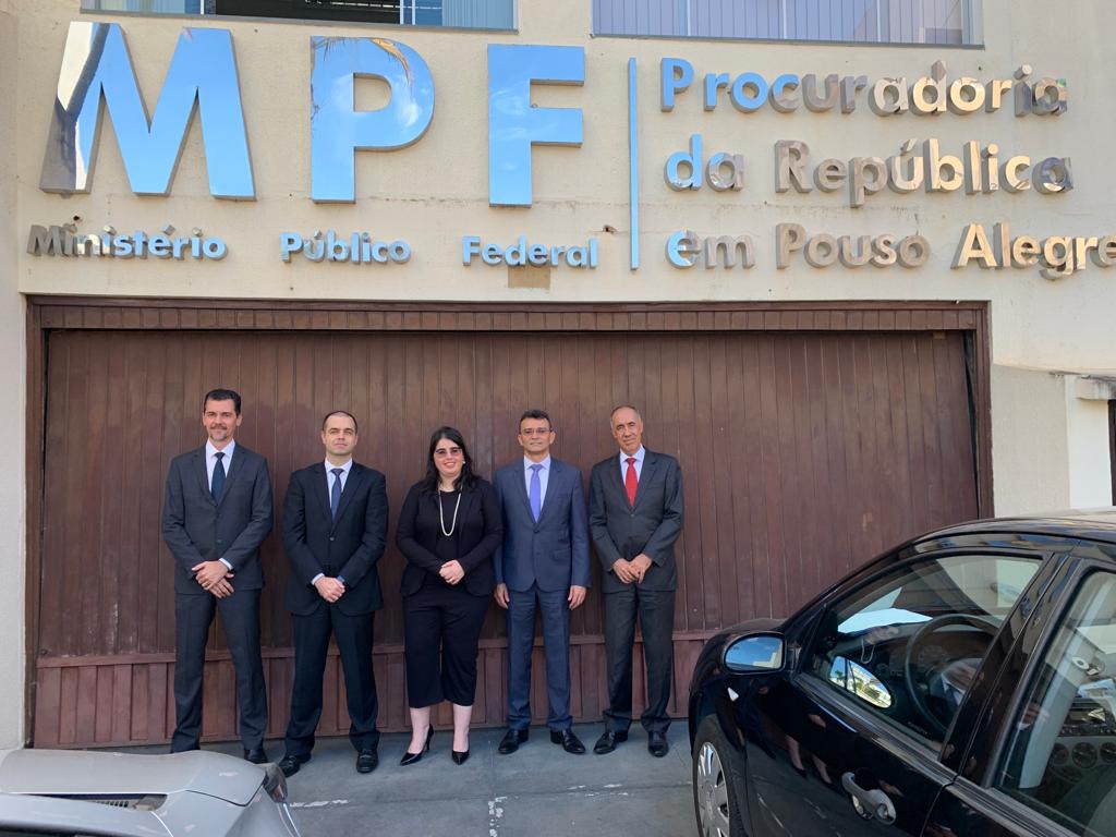 Fotografia colorida com quatro homens e uma mulher ao centro, todos com trajes formais, em frente a um portão de garagem marrom. Ao fundo, as palavras MPF - Ministério Público Federal | Procuradoria da República em Pouso Alegre.