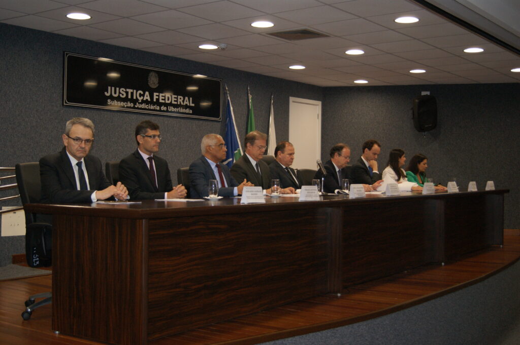 Foro colorida retangular da mesa de autoridades. Na foto, as pessoas aparecem sentadas, em trajes formais. 