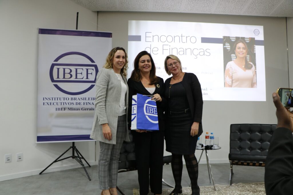 Foto colorida horizontal mostra três mulheres em pé posando para a foto no auditório. A Presidente Mônica S. segura uma sacola. 