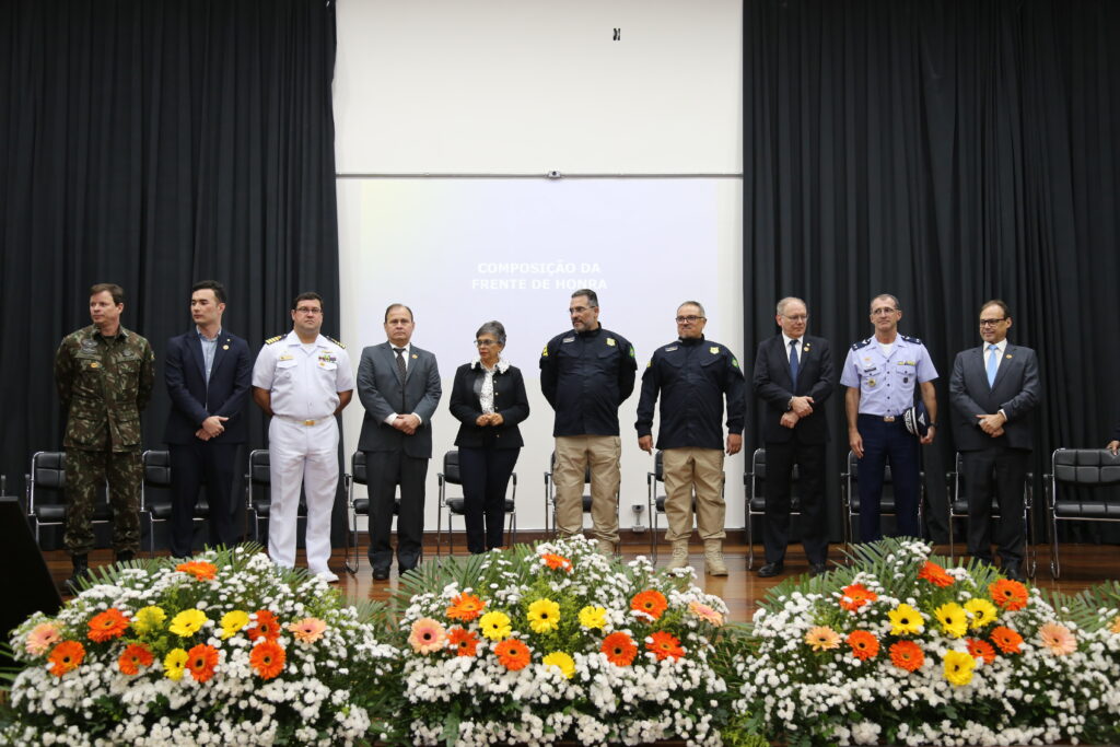 Foto colorida horizontal. Um grupo de pessoas, uma mulher e nove homens, em trajes formais está em pé no palco do evento.