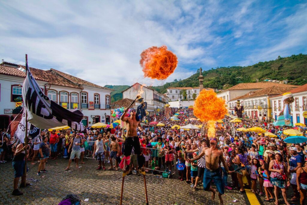 Fotografia colorida quadrada em que uma multidão de pessoas assistem a uma apresentação de malabaristas cuspindo fogo na praça Tiradentes, em Ouro Preto.