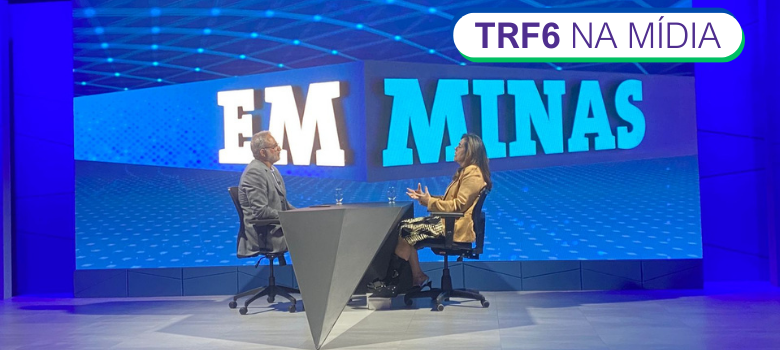 Fotografia colorida de uma mulher falando com um homem separados por uma mesa retangular. Ao fundo, marca do programa 'Em Minas' em fundo azul.