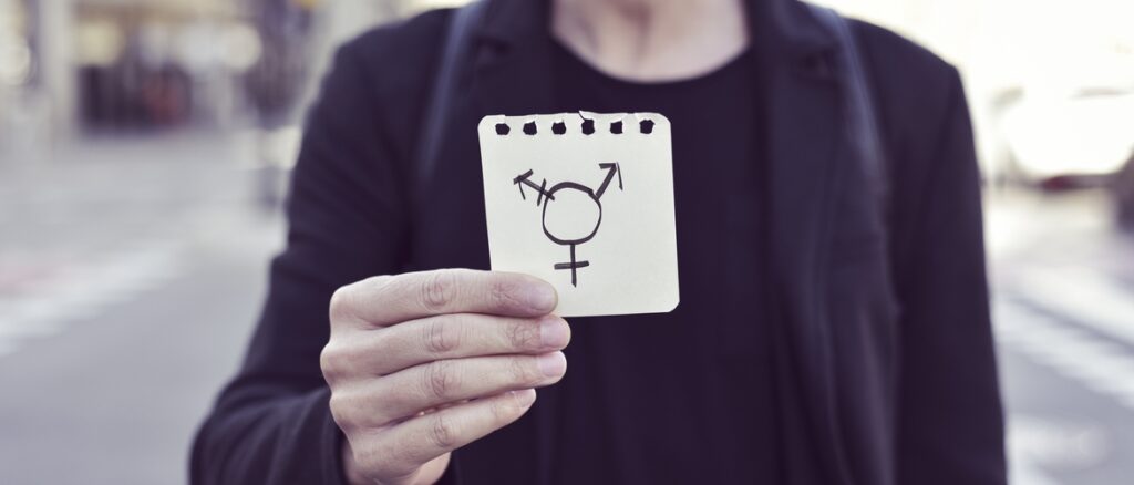 Fotografia colorida e retangular em que uma pessoa sem cabeça segura um pedaço de papel com o símbolo da transexualidade.